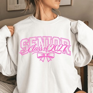 May include: A white crewneck sweatshirt featuring a bright pink graphic print on the chest. The text reads SENIOR class of 2027 in a mix of block and script fonts, accompanied by a pink bow illustration centered below the lettering.