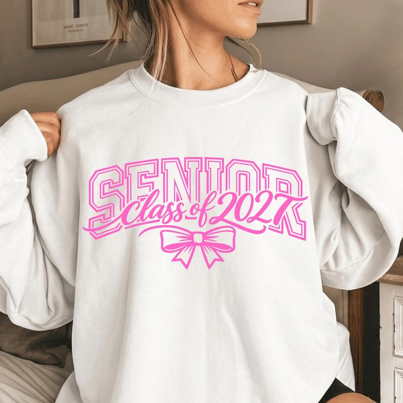 May include: A white crewneck sweatshirt featuring a bright pink graphic print on the chest. The text reads SENIOR class of 2027 in a mix of block and script fonts, accompanied by a pink bow illustration centered below the lettering.