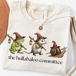Op de afbeelding: Een crèmekleurig t-shirt met een afbeelding van een kikker, een opossum en een slak, elk met een bordeauxrode tovenaarshoed op en een lantaarn aan een houten staf. De tekst onder de illustratie luidt, the hullabaloo committee.