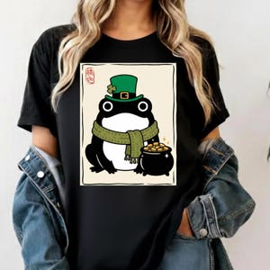 Op de afbeelding: Zwart T-shirt met een cartoonkikker met een groene hoge hoed met een klavertje vier, een groene sjaal en een pot goud. De kikker staat tegen een crèmekleurige achtergrond met een zwarte rand. Een casual kledingstuk.