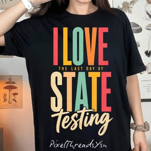 Puede incluir: Camiseta negra con el texto "I LOVE THE LAST DAY OF STATE TESTING" en letras de bloque coloridas y una fuente cursiva. La camiseta también tiene el texto "PixelThreadsYsn" en una fuente cursiva en la parte inferior.