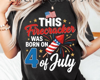 Op de afbeelding: Een zwart t-shirt met een afbeelding met de tekst THIS Firecracker WAS BORN ON 4 of July. Het ontwerp bevat een rood, wit en blauwe vuurwerkraket, een kleine Amerikaanse vlag en gele sterren op een zwarte achtergrond met rode en blauwe accenten.