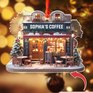 May include: A decorative Christmas ornament shaped like a miniature coffee shop. The shop has a red awning, a blue facade, and the text "SOPHIA'S COFFEE" above the entrance. The ornament includes small tables, chairs, and a Christmas tree. The text "NOTE: THIS ORNAMENT IS 2D, NOT 3D" is also present.