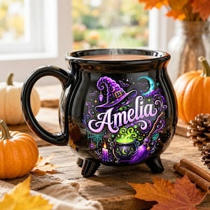 May include: A black ceramic mug shaped like a cauldron with three small feet. The front features a colorful graphic of a purple witch hat, a bubbling green potion, a broom, and the name Amelia in white script, surrounded by stars and moons.