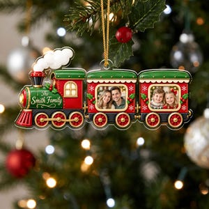 May include: A Christmas tree ornament shaped like a train with a green engine, red accents, and two passenger cars. The engine features the text The Smith Family. The two passenger cars contain photo frames displaying images of people.