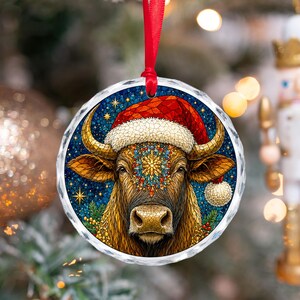 May include: A round, clear glass Christmas ornament featuring a stained-glass style illustration of a brown bull wearing a red Santa hat with white trim. The bull has a colorful, intricate floral pattern on its forehead and is set against a dark blue starry background with green pine branches and red berries.