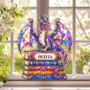 May include: A stained glass style hanging ornament featuring a purple and blue dragon perched atop a stack of five colorful books. The top book features a decorative label with the name OLIVIA printed in dark blue capital letters.