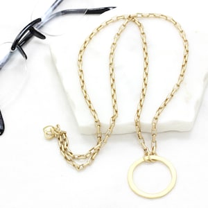 May include: A gold chain necklace with a large circle pendant. The chain is made of small, square links.
