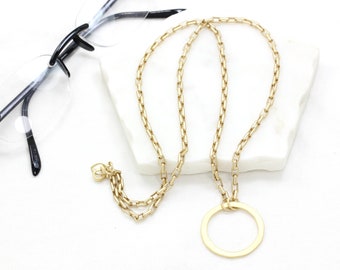 May include: A gold chain necklace with a large circle pendant. The chain is made of small, square links.