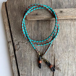 May include: A braided leather eyeglass chain with turquoise and brown accents. The chain has metal clasps and is designed to hold glasses.