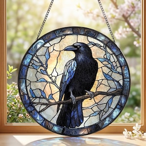 Puede incluir: Vidriera redonda con un cuervo negro posado en una rama. El cuervo tiene detalles azules y blancos en sus alas. El fondo es una mezcla de azul, amarillo y blanco, con un borde azul oscuro y una cadena para colgar.
