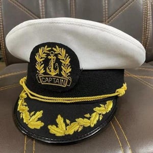 Könnte beinhalten: Eine weiße und schwarze Kapitänsmütze mit goldener Stickerei. Die Mütze hat ein goldenes Ankeremblem und das Wort "CAPTAIN". Der Rand ist mit goldenen Blattmotiven und einer goldenen Kordel verziert.
