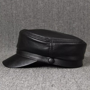 May include: A black leather cap with a short brim and a button detail. The cap has a structured design and a band around the crown. The hat is made of a smooth, shiny material.