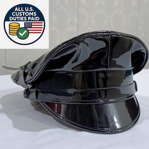 Op de afbeelding: Een glanzende zwarte hoed met een vizier en een riem. De hoed heeft een glanzende, lakleren uitstraling. De afbeelding bevat ook een cirkelvormige afbeelding met de tekst "ALL U.S. CUSTOMS DUTIES PAID".