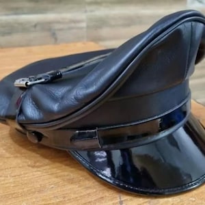 Könnte beinhalten: Ein schwarzer Lederhut mit glänzendem Lackleder-Schirm. Der Hut hat eine abgerundete Krone und einen Riemen mit einer silbernen Schnalle. Der Hut liegt auf einer Holzoberfläche.