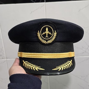 May include: A navy blue pilot's hat with gold embroidery. The hat has a gold emblem with a plane inside a wreath, gold bands, and leaf designs. The hat is held by a person.