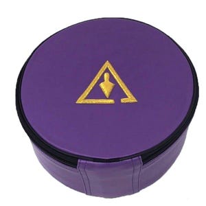Op de afbeelding: Een ronde, paarse koffer met een zwarte ritssluiting en een goud geborduurd symbool op de bovenkant. Het symbool is een driehoek met een figuur erin. De koffer lijkt gemaakt van een leerachtig materiaal.