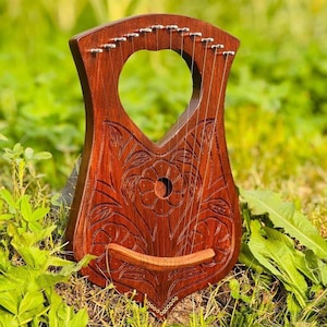 May include: A dark brown wooden lyre featuring a heart-shaped sound hole at the top and intricate floral carvings across the front body. The stringed musical instrument includes a horizontal bridge and metal tuning pins along the upper edge.