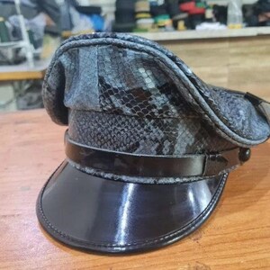 Könnte beinhalten: Ein grau-schwarz gemusterter Hut mit schwarzem Schirm und Band. Der Hut hat ein strukturiertes Design und einen glänzenden, lacklederartigen Schirm. Der Hut liegt auf einer Holzoberfläche.