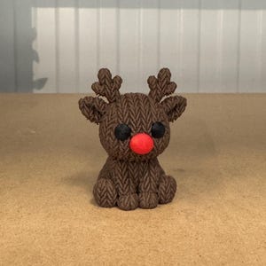 以下が含まれることがあります： 織り目模様の小さな茶色のトナカイの置物。トナカイは黒い目と明るい赤色の鼻をしています。角も茶色です。置物は薄茶色の表面に置かれています。