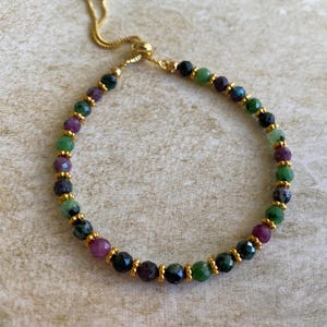 May include: A bracelet with a mix of green, purple, and black gemstones, with small gold-coloured beads. The bracelet has a gold-coloured chain and clasp for adjustable sizing. The gemstones are round and vary in size.