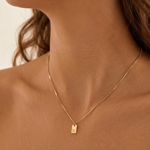 Op de afbeelding: Een goudkleurige ketting met een fijne boxketting en een kleine, rechthoekige hanger. De hanger is gegraveerd met de hoofdletter R in een schreeflettertype. De ketting rust op de huid en heeft een gepolijste, metalen afwerking.