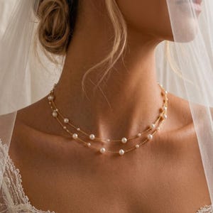 May include: A delicate double-strand necklace featuring small, round white pearls spaced evenly along a thin gold chain. The necklace rests against the neck, showcasing a layered design with a polished metallic finish and classic pearl accents.