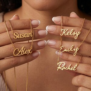 Pode incluir: Colares dourados com os nomes "Susan", "Alex", "Kelsy", "Emily" e "Rachel". Os colares são exibidos em um fundo neutro, mostrando a escrita delicada e os detalhes da corrente. Uma joia personalizada.
