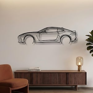 Puede incluir: Una pieza de arte mural de metal negro que representa el perfil lateral de un coche deportivo. La obra de arte está montada en una pared blanca sobre un mueble de madera. Una planta en maceta y una silla moderna están en primer plano.