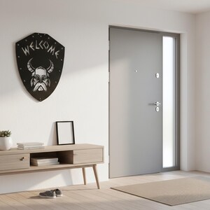 Op de afbeelding: Een zwart metalen wandbord in de vorm van een schild met een afbeelding van een gehoornde Vikinghelm en het woord WELCOME in een gestileerd lettertype. Het bord is bevestigd aan een witte muur boven een console van licht hout met lades.