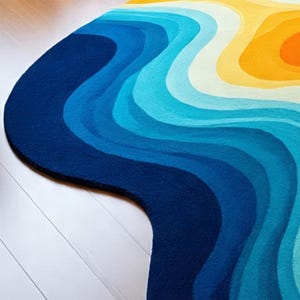 May include: A rug with a wavy design in shades of blue, white, yellow, and orange. The rug's pattern resembles a topographic map or a stylized ocean scene. The rug is placed on a white wooden floor.
