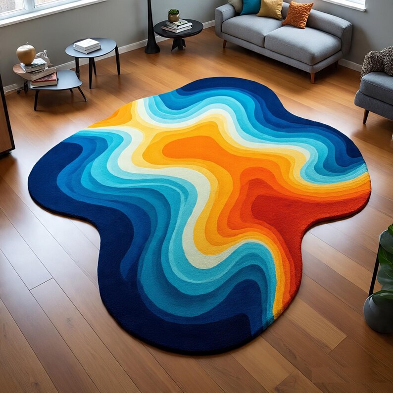 May include: A large, abstract area rug with a unique, organic shape. The rug features a gradient design with bands of color, including shades of blue, orange, yellow, and red. The rug is placed on a hardwood floor in a living room setting.
