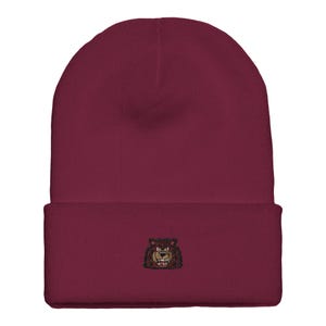 Puede incluir: Un gorro granate con borde doblado. El gorro presenta una imagen bordada de la cara de un oso pardo en la parte delantera. El oso tiene una expresión de enfado con dientes visibles y un contorno oscuro.