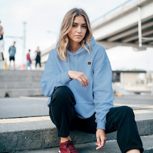 Puede incluir: Una sudadera con capucha azul claro con un pequeño diseño bordado marrón en el pecho. La sudadera con capucha se combina con pantalones negros y zapatos burdeos. La persona está sentada en escalones de hormigón al aire libre.