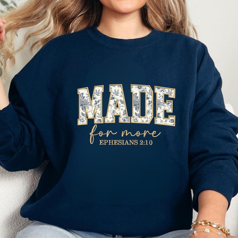 May include: Navy blue sweatshirt with the words "MADE for more" in a floral design. The text "Ephesians 2:10" is below the main text. The sweatshirt has a crew neck and long sleeves.