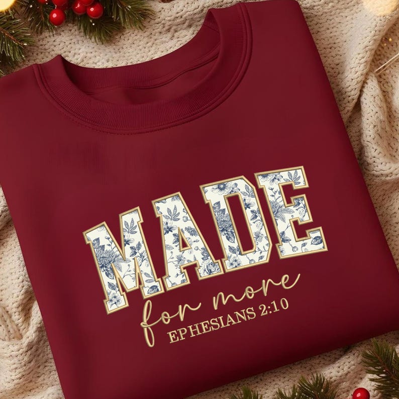 May include: A maroon sweatshirt with the words "MADE" in large, gold-outlined letters filled with a blue and white floral pattern. Below, in cursive, it says "for more" and "EPHESIANS 2:10".