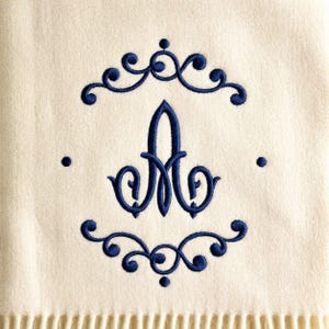 以下が含まれることがあります： クリーム色の生地に、濃い青色の糸で刺繍された文字「A」のモノグラム。文字は、対称的な渦巻きとドットで構成された装飾的なフレームの中央に配置されており、すべて濃い青色の糸で生地の背景に縫い付けられています。