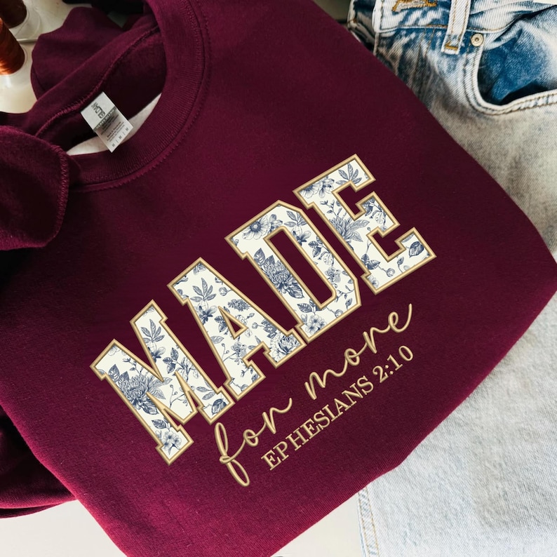 May include: Burgundy sweatshirt with the word "MADE" in a floral design, outlined in gold. The phrase "for more" and "Ephesians 2:10" are written in gold script. The sweatshirt is folded, with a pair of jeans in the background.