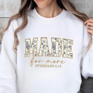 May include: White sweatshirt with the words "MADE" in a gold outline, filled with a blue floral pattern. Below, the words "for more" are written in a cursive font, with "EPHESIANS 2:10" below.
