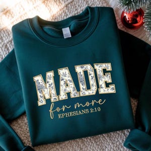 May include: A dark teal sweatshirt with the word "MADE" in large, block letters filled with a floral pattern. Below, the phrase "for more" is written in a cursive font, with "EPHESIANS 2:10" underneath. The sweatshirt is folded.