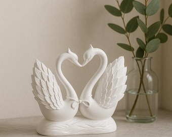 May include: A white ceramic swan figurine with two swans facing each other, forming a heart shape with their necks. The swans have detailed feather wings and are tied together with a small bow. The figurine is on a light gray surface.
