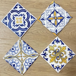Könnte beinhalten: Vier quadratische Keramikfliesen mit aufwendigen blau-gelben Blumenmustern auf weißem Grund. Die Fliesen sind quadratisch angeordnet und zeigen die dekorativen Designs. Die Fliesen liegen auf einer Holzoberfläche.