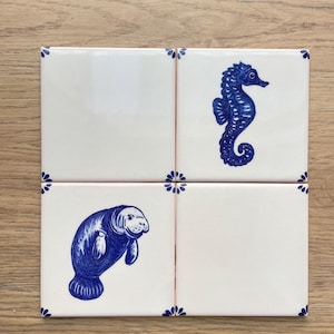 Op de afbeelding: Vier vierkante keramische tegels met een witte achtergrond en blauwe decoratieve accenten. E&eacute;n tegel bevat een illustratie van een blauw zeepaardje en een andere tegel toont een illustratie van een blauwe lamantijn. Elke hoek heeft een klein blauw bloemmotief.