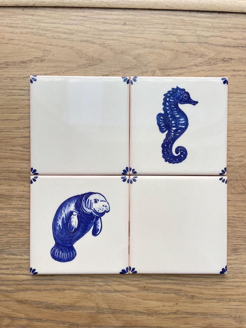 Op de afbeelding: Vier vierkante keramische tegels met een witte achtergrond en blauwe decoratieve accenten. E&eacute;n tegel bevat een illustratie van een blauw zeepaardje en een andere tegel toont een illustratie van een blauwe lamantijn. Elke hoek heeft een klein blauw bloemmotief.