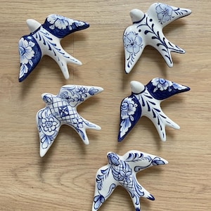May include: Five ceramic swallow figurines in white and blue. Each bird has a white head and is decorated with blue floral and geometric patterns. The swallows are arranged on a light brown wooden surface.