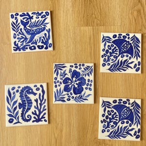 Könnte beinhalten: Fünf quadratische Keramikfliesen mit handgemalten blau-weißen Pflanzen- und Meeresmotiven. Die Fliesen zeigen einen Wal, ein Seepferdchen, eine Hibiskusblüte und zwei Rochen, umgeben von detaillierten Blatt- und Korallenmustern.