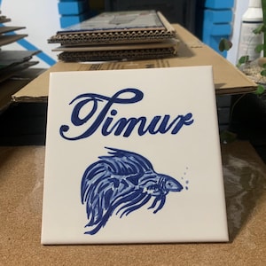 Op de afbeelding: Een vierkante witte keramische tegel met de naam Timur in een donkerblauw cursief handschrift. Onder de tekst staat een handgeschilderde illustratie van een kempvis in donkerblauwe tinten, met lange vloeiende vinnen en kleine belletjes.
