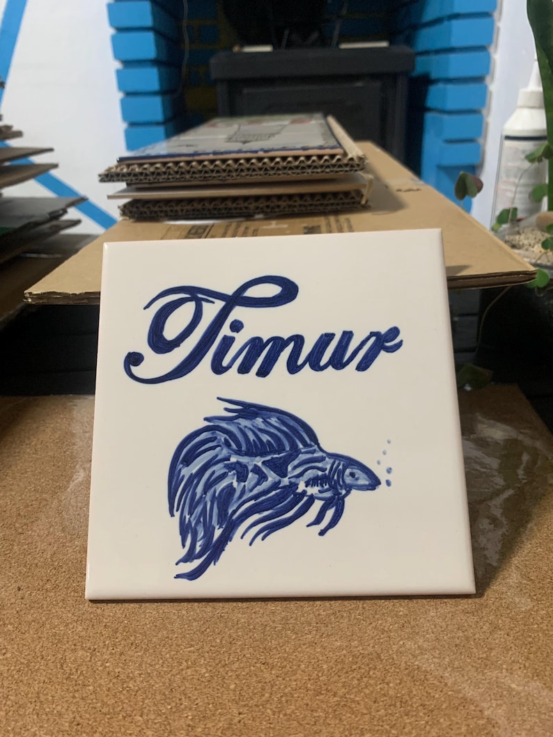 Op de afbeelding: Een vierkante witte keramische tegel met de naam Timur in een donkerblauw cursief handschrift. Onder de tekst staat een handgeschilderde illustratie van een kempvis in donkerblauwe tinten, met lange vloeiende vinnen en kleine belletjes.