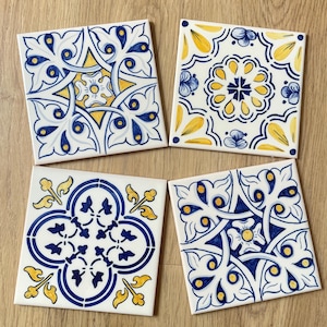 May include: Four square ceramic coasters with intricate blue and yellow floral patterns on a white background. Each coaster features a unique design, creating a set with a cohesive aesthetic. The coasters are arranged on a wooden surface.