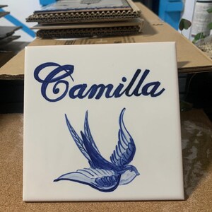 Op de afbeelding: Een vierkante witte keramische tegel met de naam Camilla in een donkerblauw cursief handschrift. Onder de naam staat een handgeschilderde illustratie van een zwaluw in blauwtinten, afgebeeld in vlucht met wijd gespreide vleugels op de witte achtergrond.